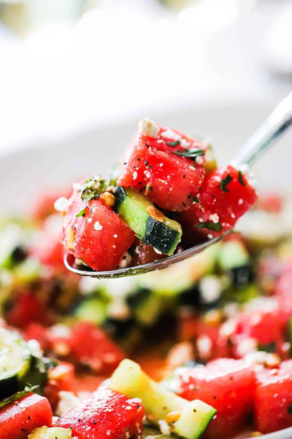 A large silver spoon lifting up a helping of watermelon cucumber salad with basil and feta.