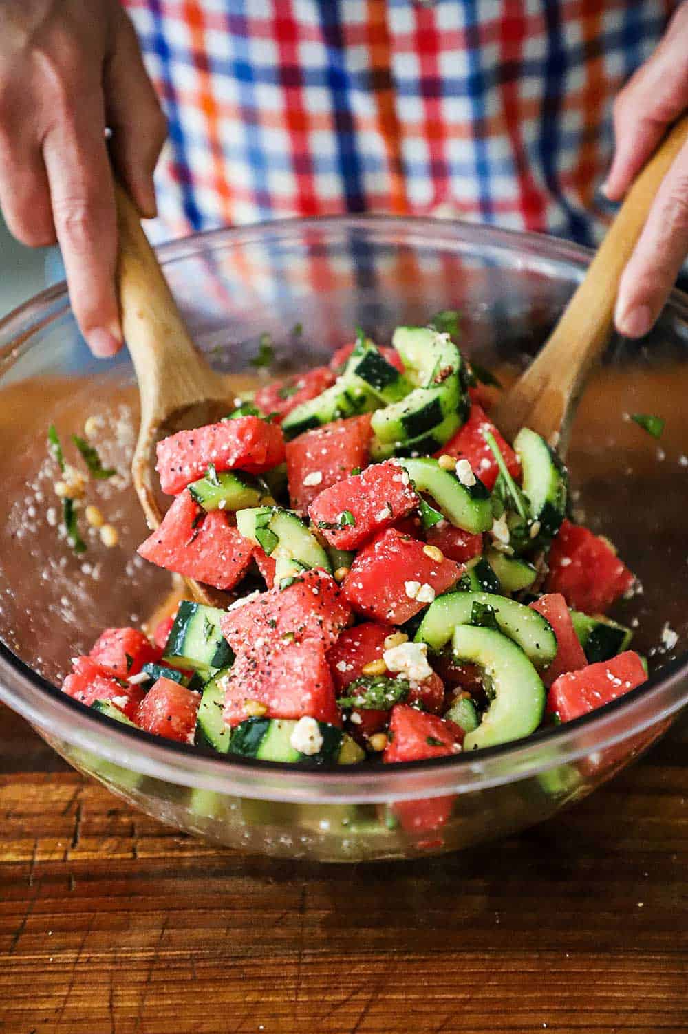 A person tossing together chunks of watermelon with sliced cucumbers, feta cheese, basil, and pine nuts in a glass bowl.