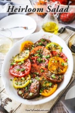 An overhead view of a large white oval platter filled with a sliced heirloom tomato salad and surrounded by a glass of white wine, olive oil, and white plate.