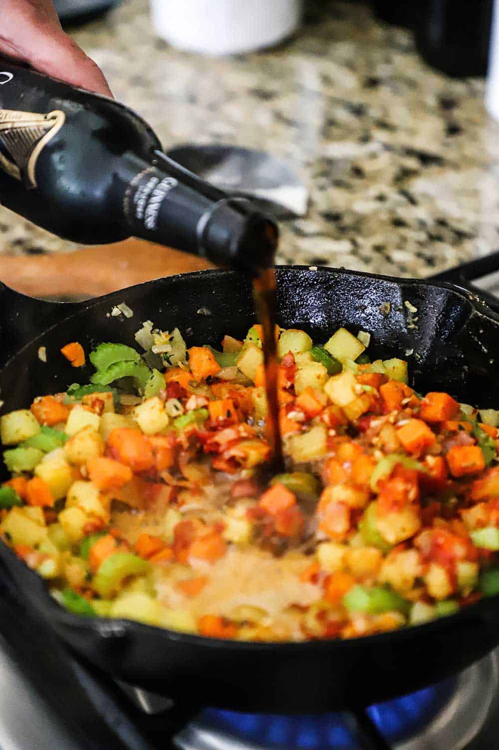 A person pouring stout beer from a bottle into a skillet filled with sautéed carrots, potatoes, and celery.