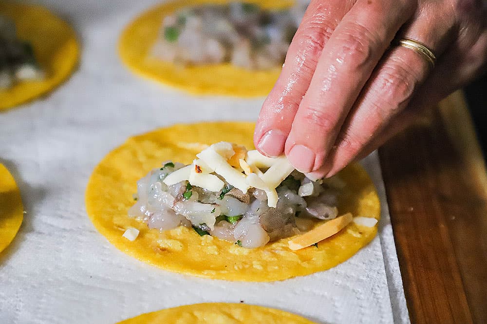 A person placing shredded cheese over uncooked chopped shrimp on an oiled corn tortilla sitting on paper towels.