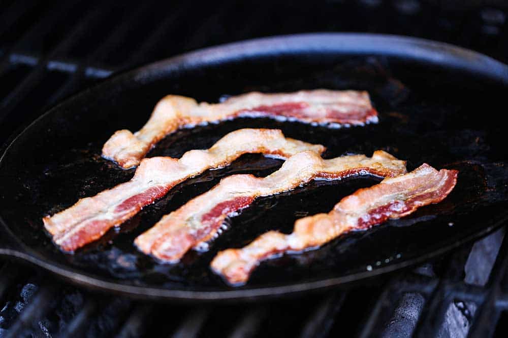 Four strips of bacon being cooked on a fajita pan on a gas grill