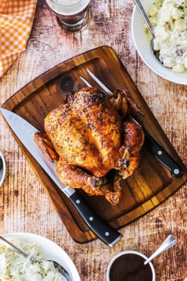 A grilled whole beer-can chicken sitting on a cutting board next to a carving knife and a bowl of BBQ sauce