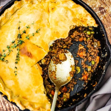 An overhead view of a Guinness ground beef pot pie in a cast iron skillet with half of the crust gone and a spoon inserted in the meat and vegetable filling.