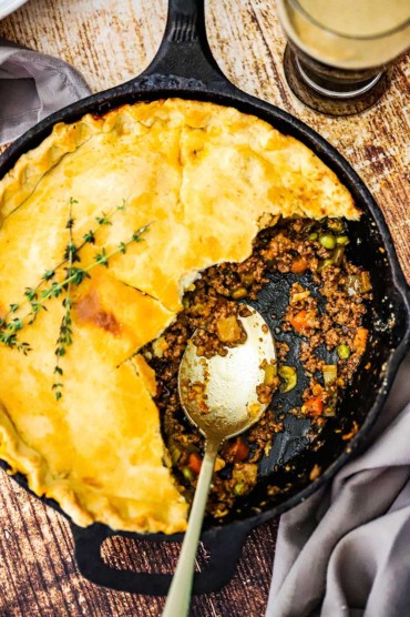 An overhead view of a Guinness ground beef pot pie in a cast iron skillet with half of the crust gone and a spoon inserted in the meat and vegetable filling.