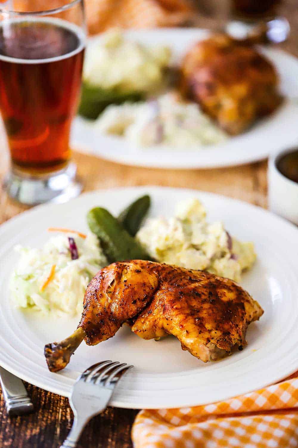 A white dinner plate filled with a beer can chicken let quarter and coleslaw, pickles, and potato salad.