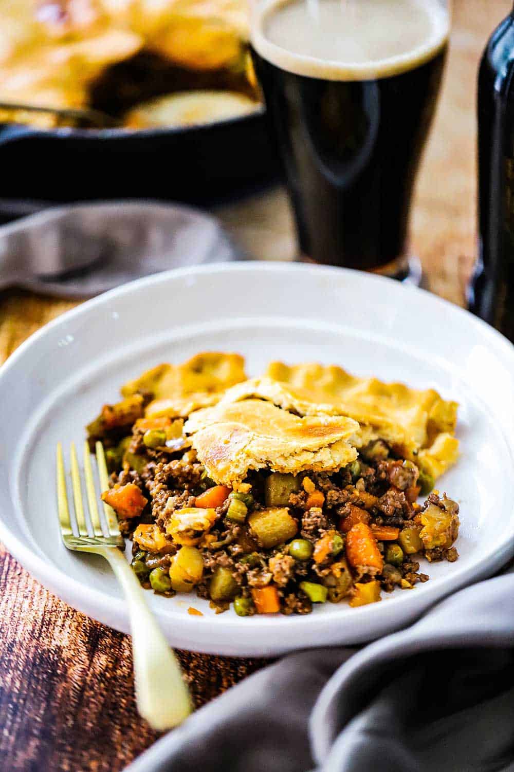 A white serving bowl filled with a serving of ground beef pot pie next to a tall glass of stout beer.