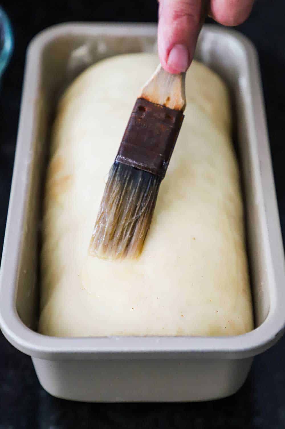 A person using a small kitchen brush to smear melted butter over the top of uncooked bread dough in a metal loaf pan.