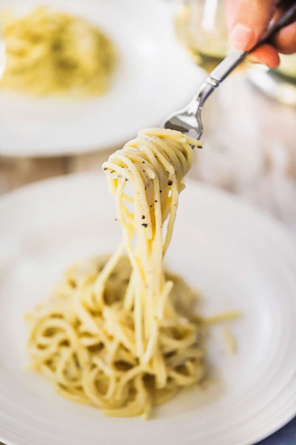 A person using a fork to lift up a helping of cacio e pepe from a white dinner plate. 