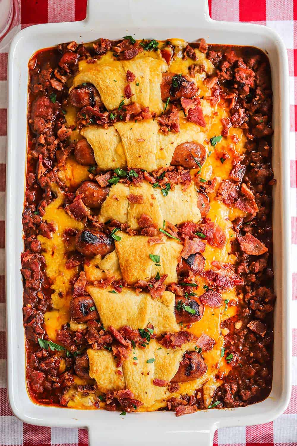 An overhead view of a chili cheese dog casserole in a white baking dish sitting on a red checkered tablecloth.