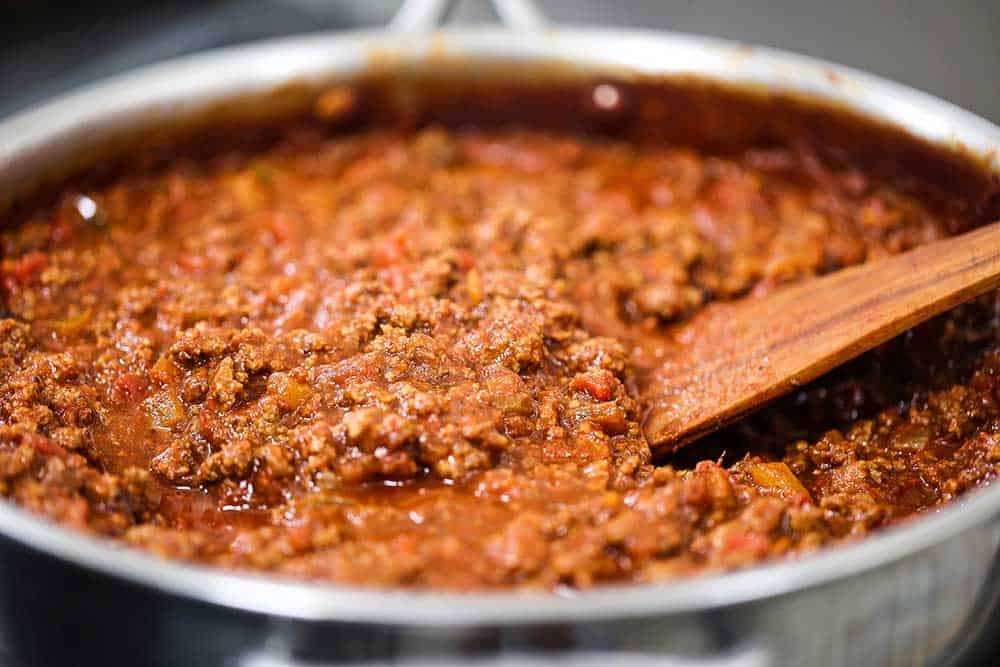 A close-up view of a large skillet filled with beef chili with a wooden spatula inserted into it.