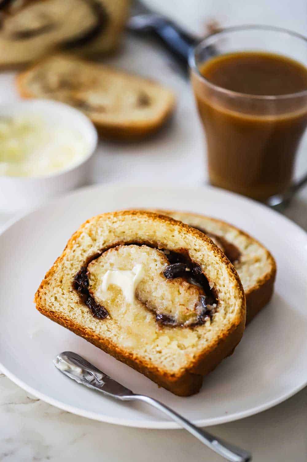 Two slices of cinnamon raisin bread stacked on top of each other with melted butter on one of the slices all on a plate next to a mug of coffee.