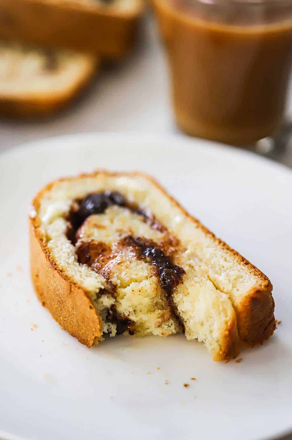 A slice of cinnamon raisin bread with a bite taken out of it sitting on a small white plate next to a mug of coffee.