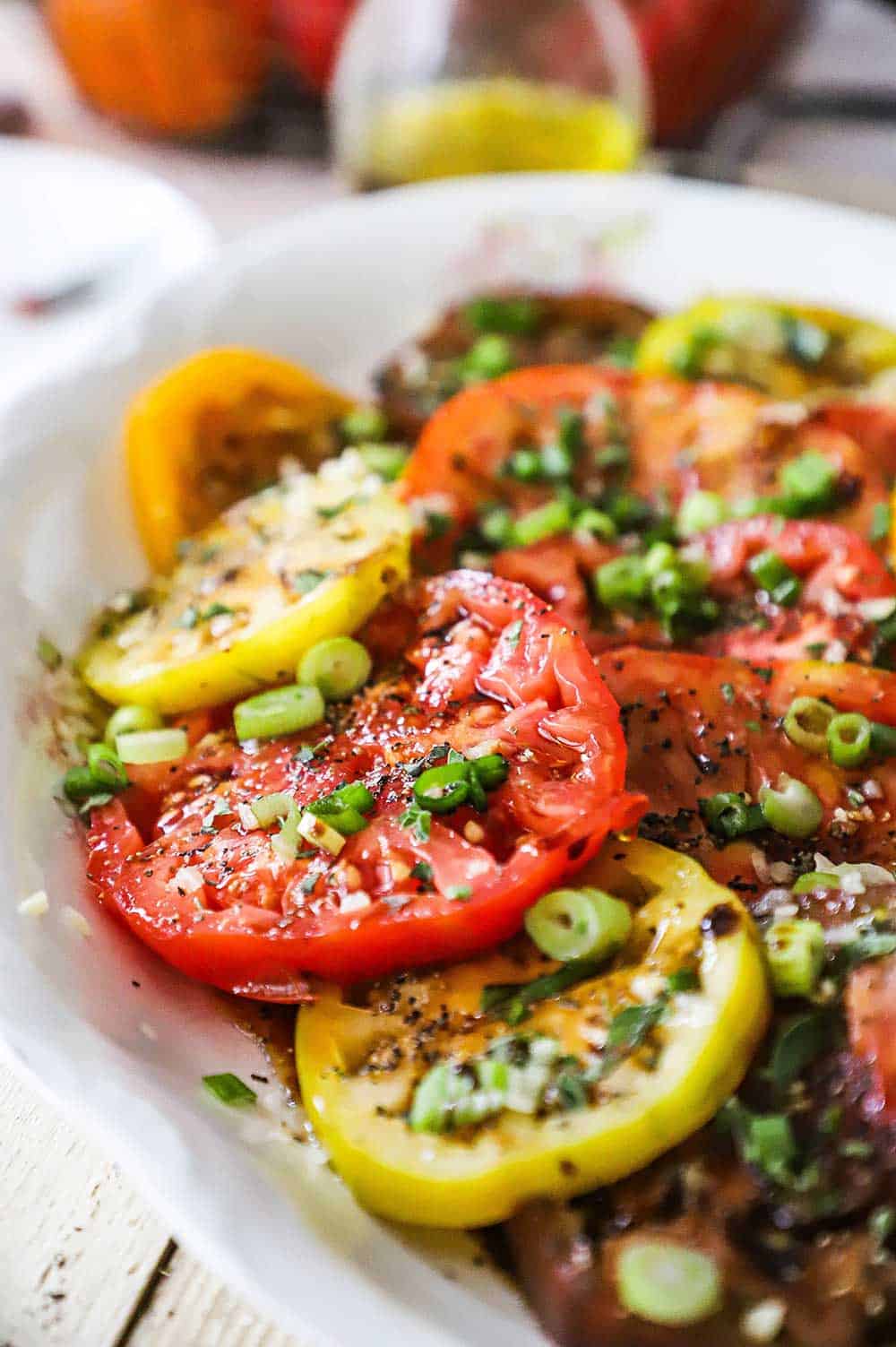 A close-up view of an heirloom tomato salad on a large oval platter topped with sliced scallions, olive oil, and balsamic vinegar.