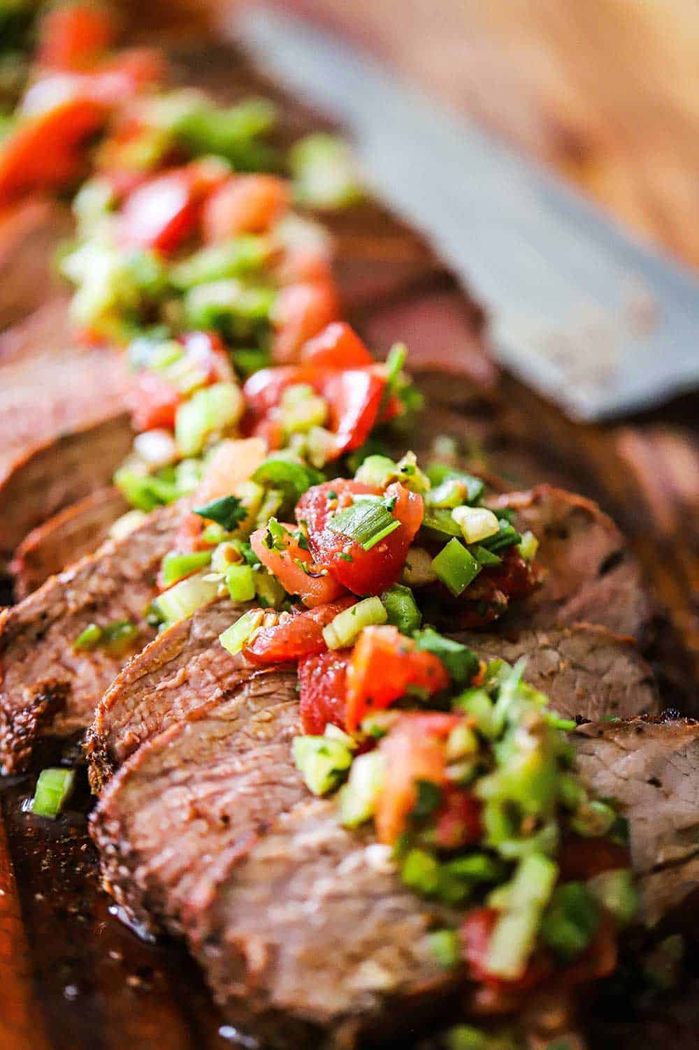 A close-up view of a grilled trip-tip with Santa Maria salsa on a cutting board next to a large knife. 