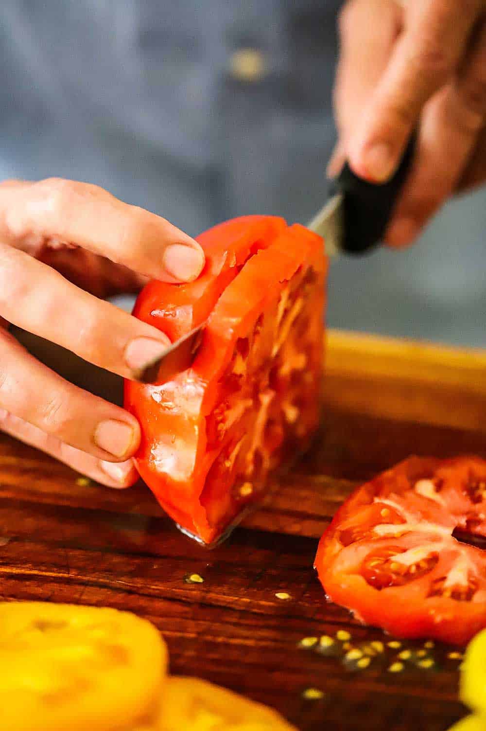 A person using a knife to cut a red heirloom tomato into slices on a wooden cutting board.