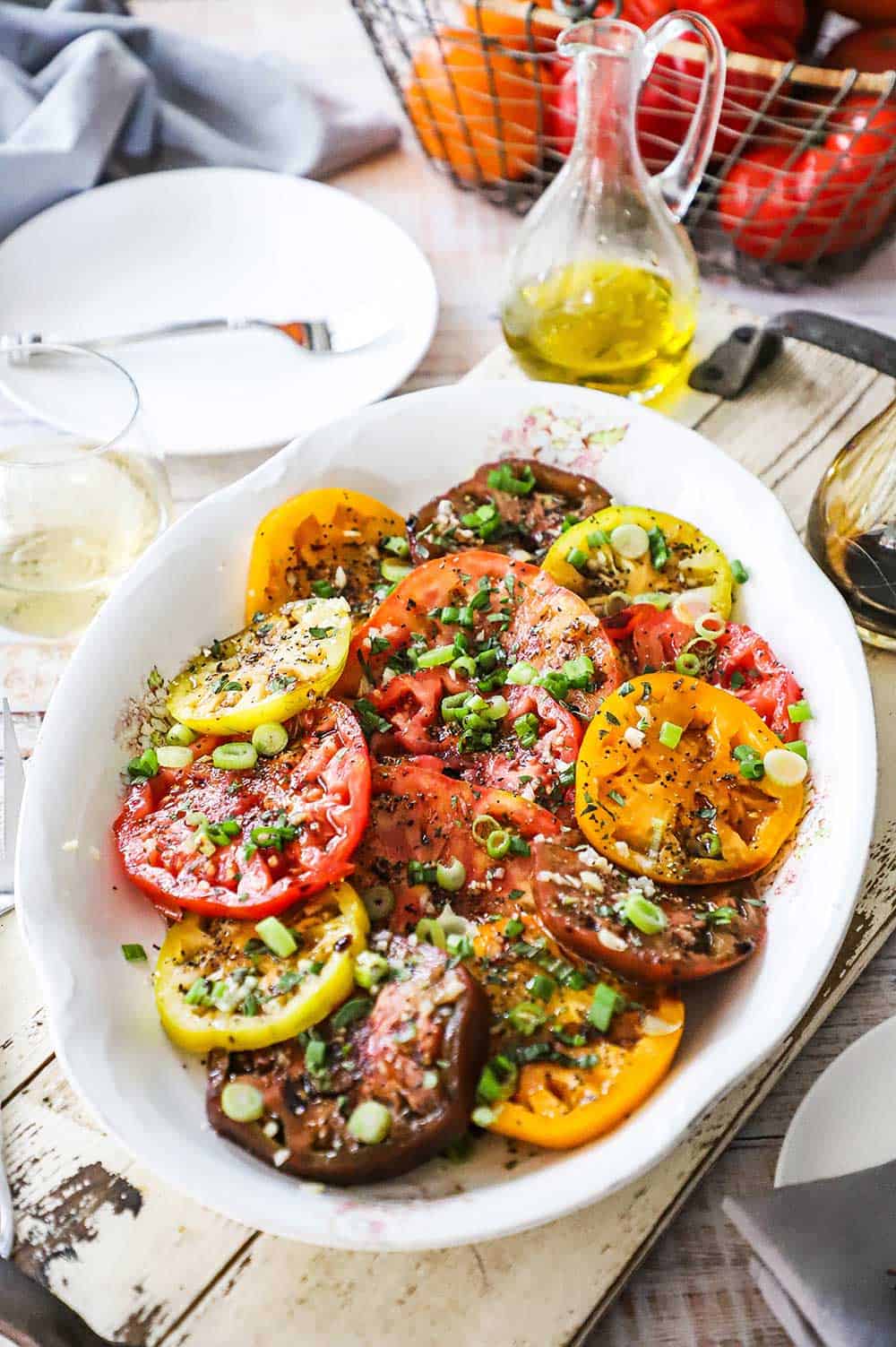 An overhead view of a large white oval platter filled with a sliced heirloom tomato salad and surrounded by a glass of white wine, olive oil, and white plate.