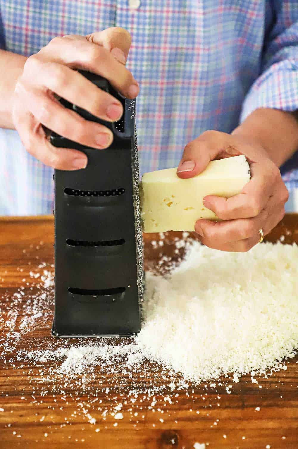 A person using a box grater to grate a block of Pecorino Romano cheese on a wooden cutting board.
