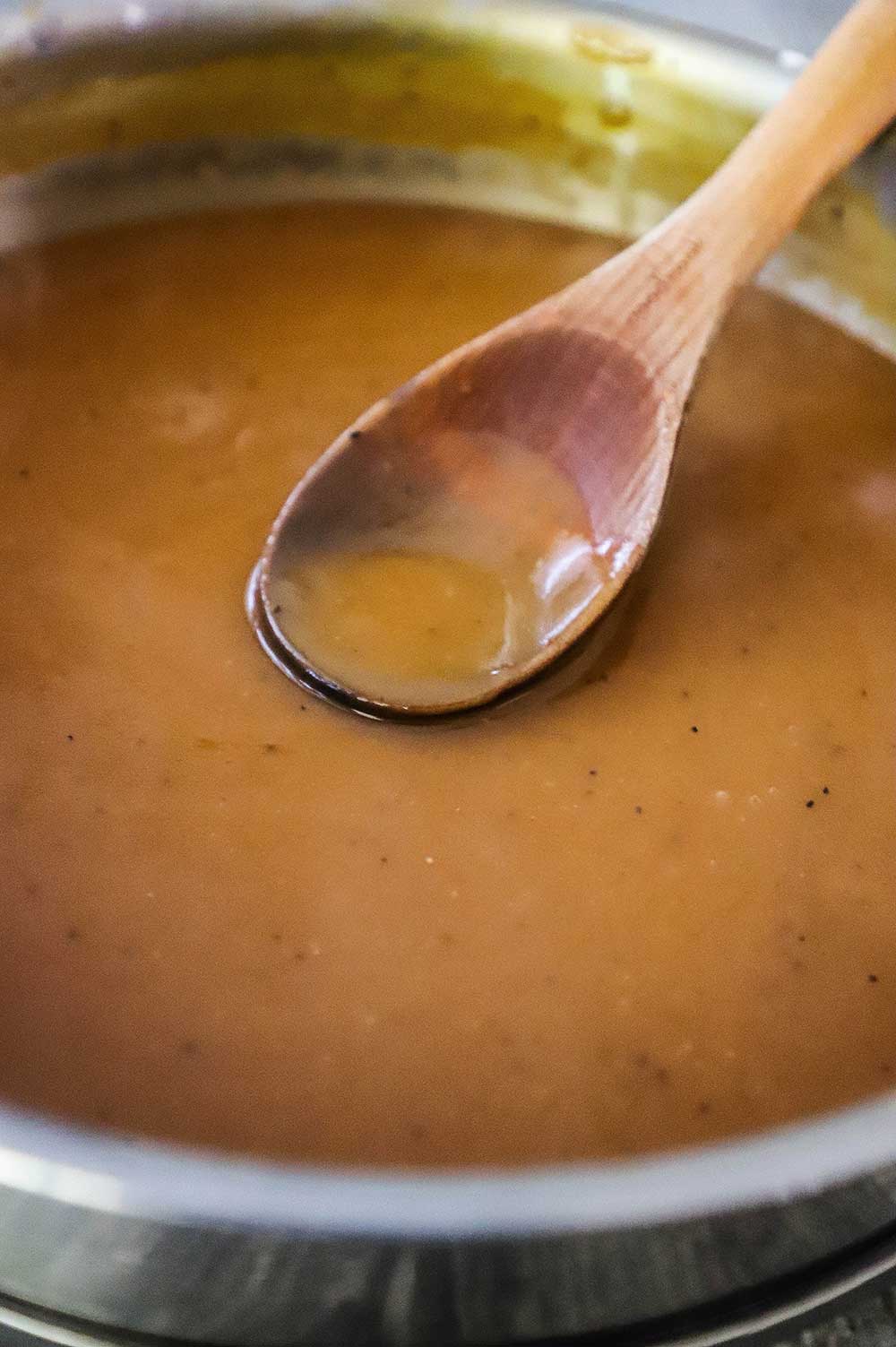 A large sliver saucepan filled with brown gravy with a wooden spoon in the middle of the pan.