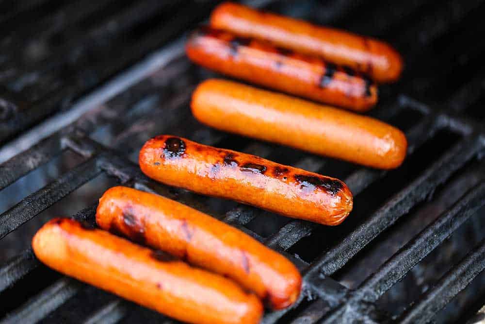 Six hot dogs being grilled on the grates of a gas grill.