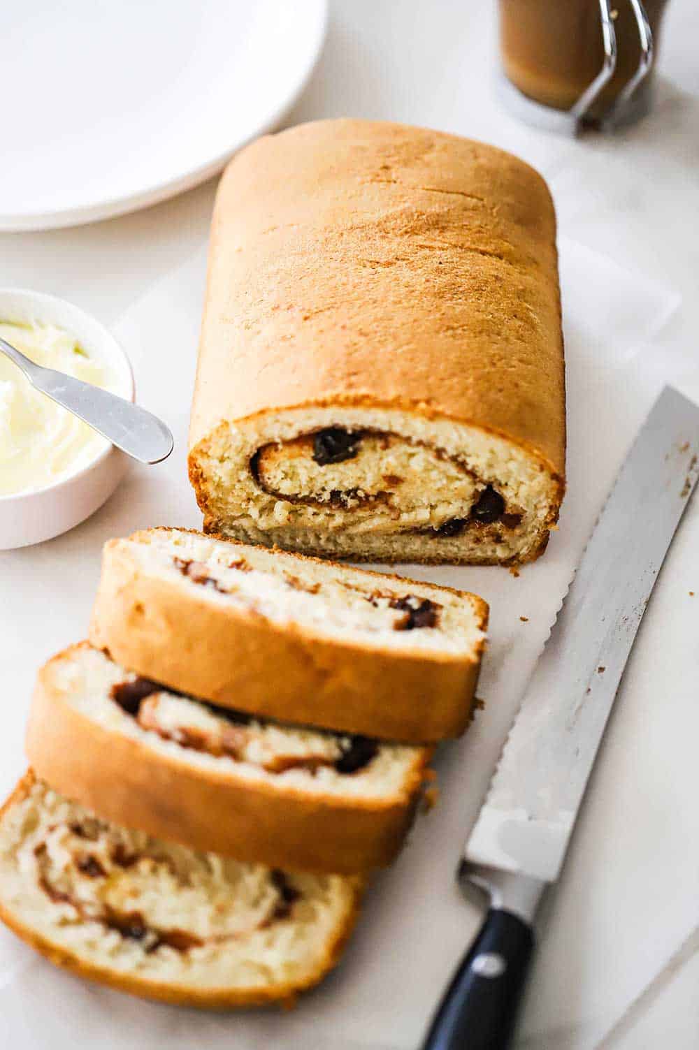 A loaf of cinnamon raisin bread that has three slices cut and are sitting on parchment pater next to a serrated knife and a bowl of softened butter.