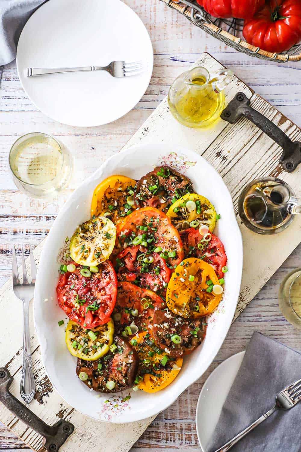 An overhead view of a platter filled with sliced tomatoes that are red, purple, and yellow and topped with scallions, olive oil, and balsamic vinegar.