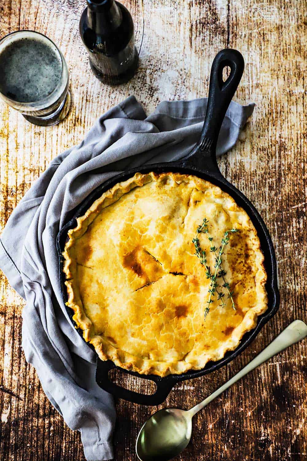 An overhead view of an untouched Guinness ground beef pot pie covered with a pie crust and sitting new to a bottle and glass of stout beer.