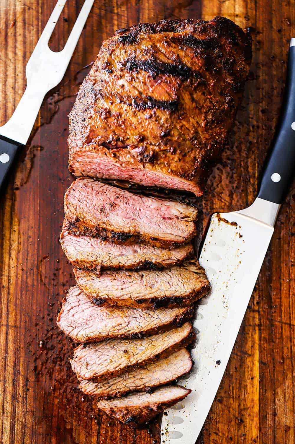 A sliced grilled tri-tip steak on a cutting board with a large knife next to it. 