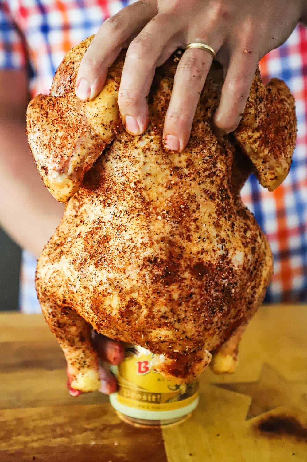 A person placing a seasoned whole uncooked chicken onto a beer can sitting on a cutting board