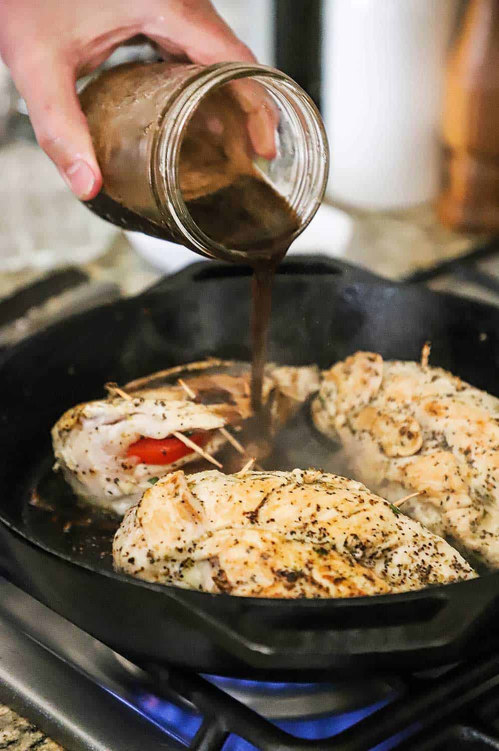 A person pouring a balsamic mixture from a small Mason jar into a skillet filled with 3 caprese-stuffed chicken breasts. 