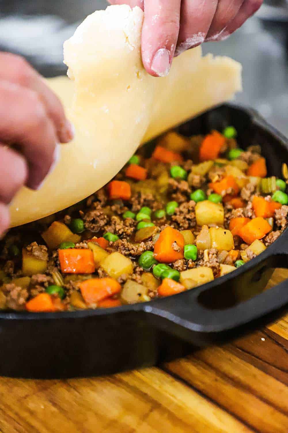 A person placing pie dough over the top of a cast-iron skillet filled with a ground beef and sautéed vegetable filling.