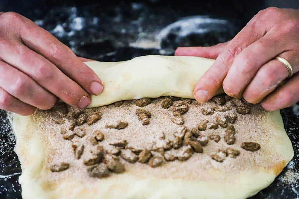 A person rolling bread dough with cinnamon and raisins in the center on a floured surface.