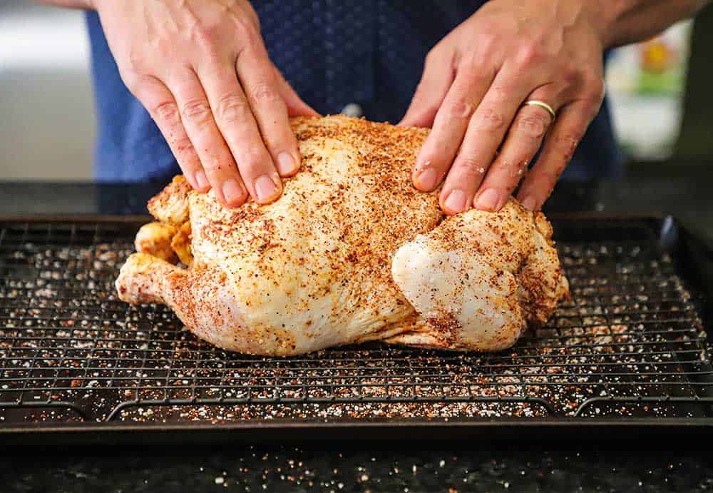 Two hands pressing barbecue rub all over a whole uncooked chicken sitting on a baking rack over a baking sheet.