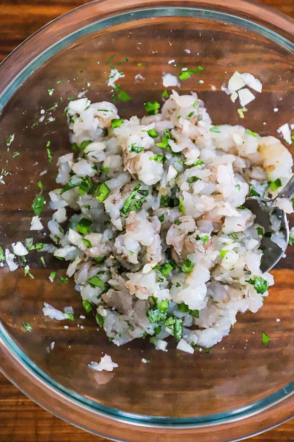 An overhead view of a glass bowl filled with roughly chopped uncooked shrimp combined with chopped cilantro and jalapeños