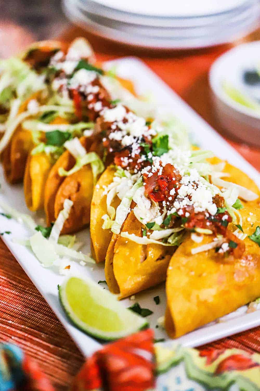 A white rectangular platter filled with shrimp tacos dorados and topped with lettuce, salta, and crumbled cheese.