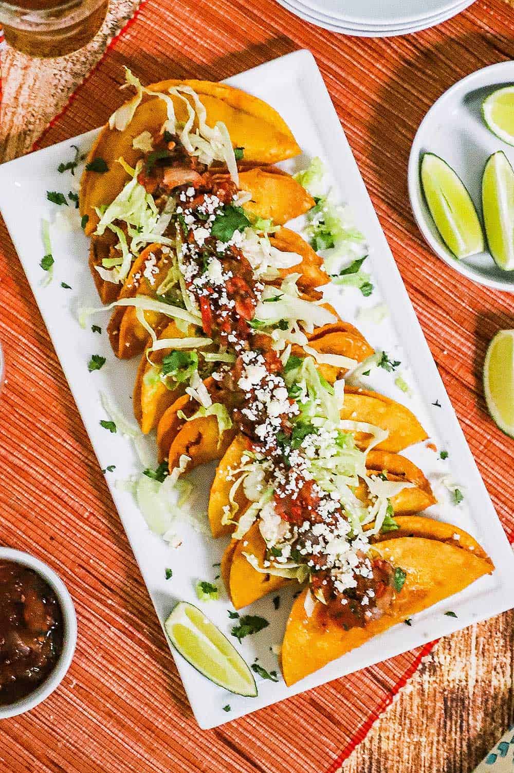 An overhead view of a white rectangular platter filled with a lineup of shrimp tacos dorados and surrounded by limes, salsa, and a glass of beer.