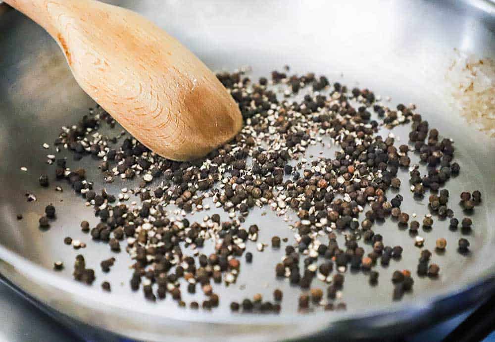Black peppercorns being toasted in a large stainless steel skillet with a wooden spoon moving them around.