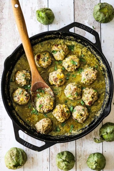 An overhead view of albondigas with salsa verde sauce in a large black cast-iron skillet with a wooden spoon in the pan.