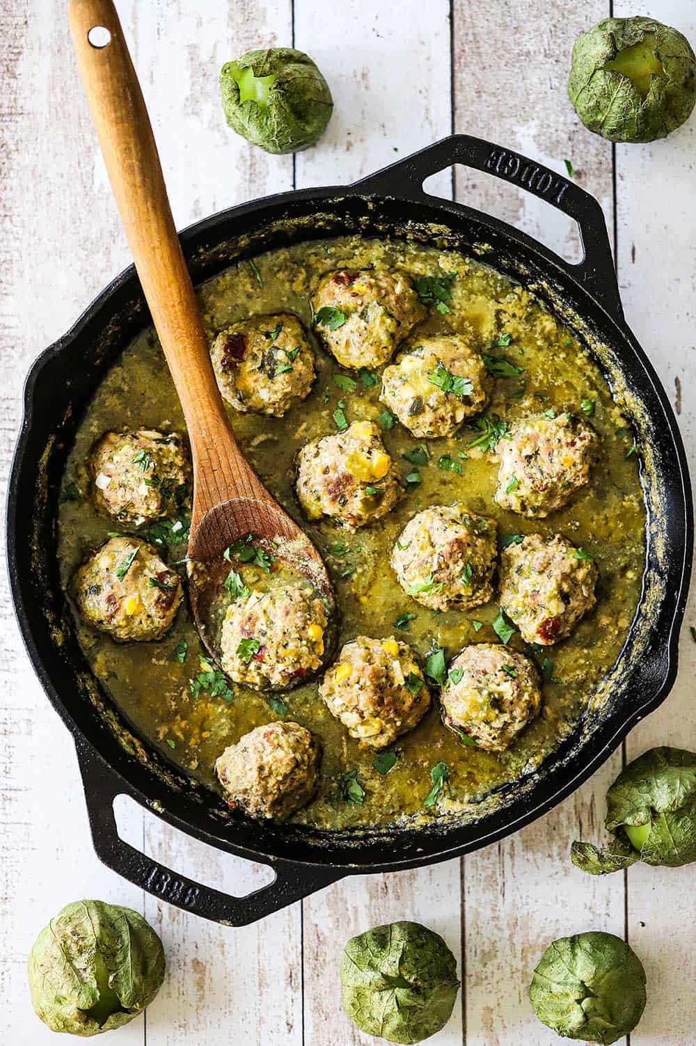 An overhead view of albondigas with salsa verde sauce in a large black cast-iron skillet with a wooden spoon in the pan.