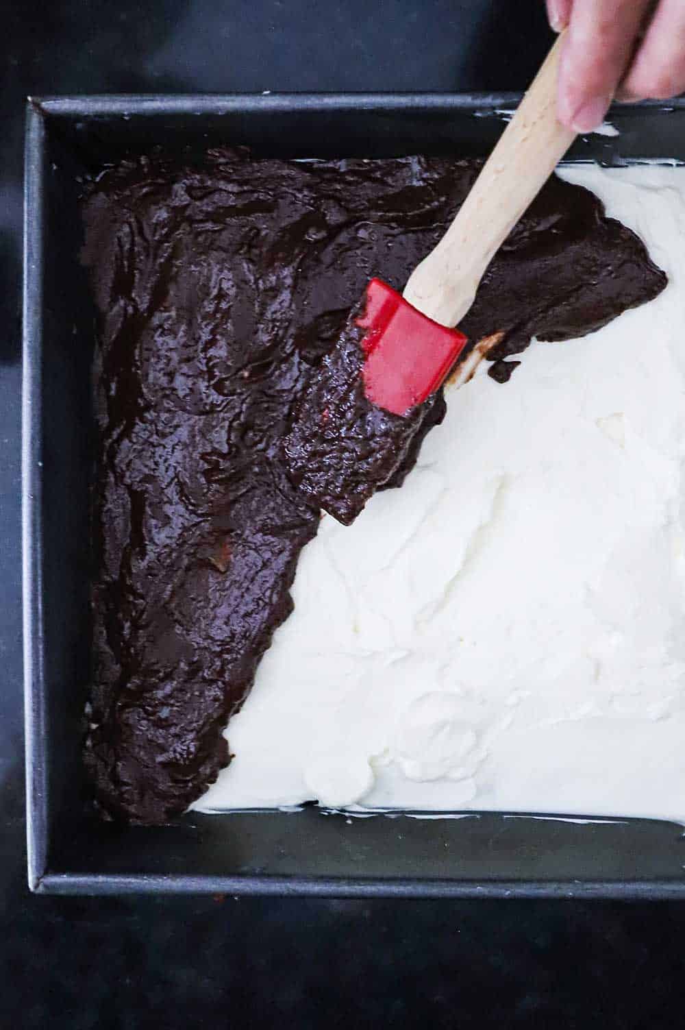 A red using a spatula to spread homemade chocolate pudding over the top of a cream cheese filling in a square metal baking pan. 