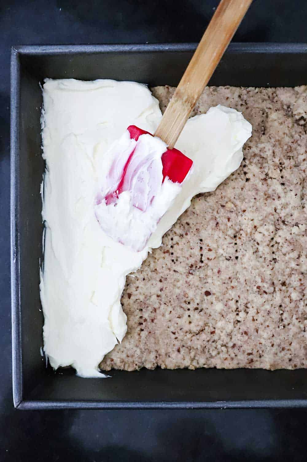 A red spatula being used to spread cream cheese filling over a short bread crust in a metal baking pan.