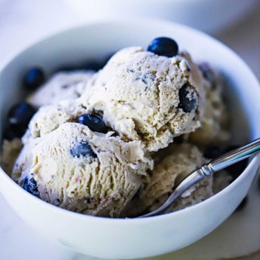 A white ice cream bowl filled with several scoops of homemade blueberry ice cream with a spoon stuck in the side.