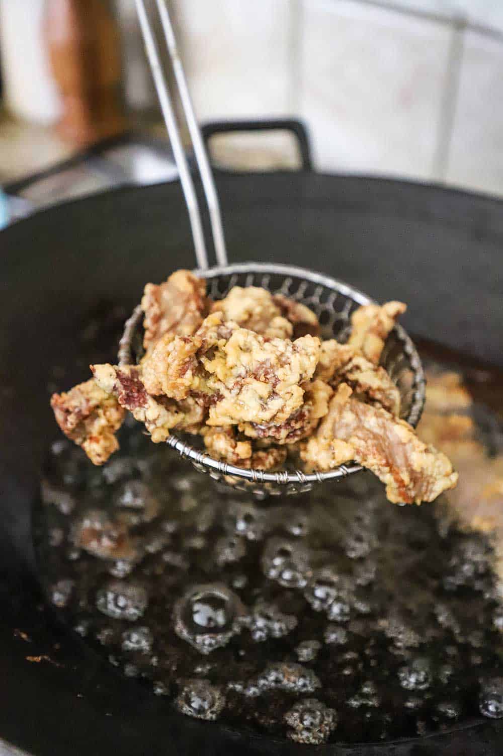 A metal spider that is holding up deep fried beef strips over a wok filled with hot oil.