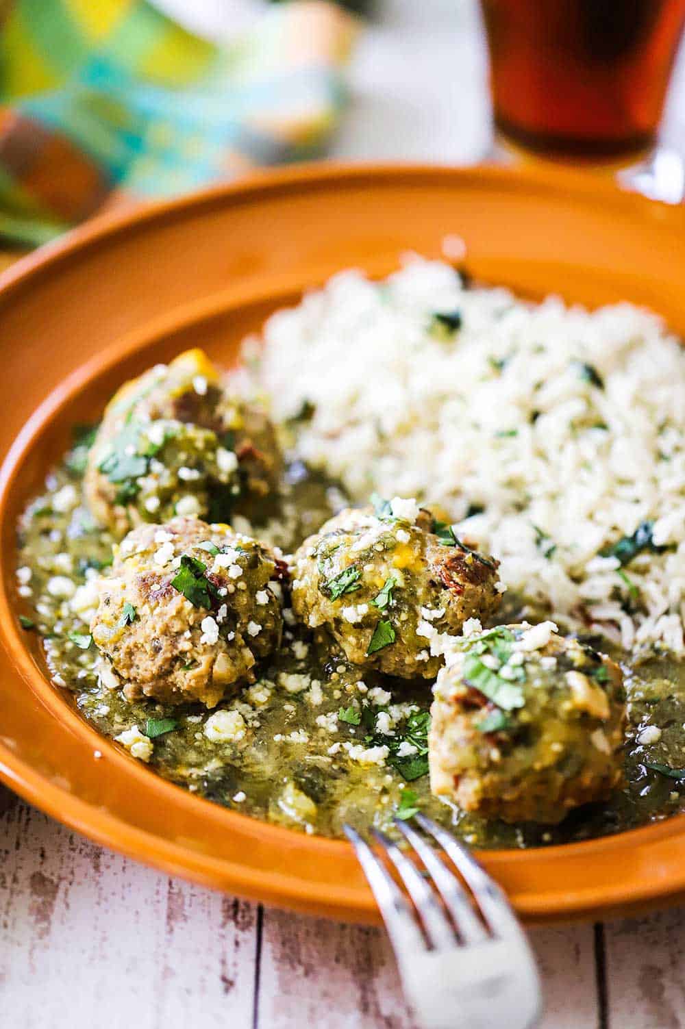 An orange dinner plate filled with four albondigas on a bed of salsa verde and a side of cilantro rice.