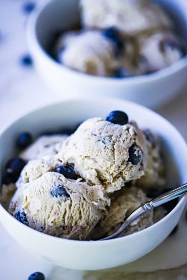 Two white ice cream bowls filled with homemade blueberry ice cream with fresh blueberries sprinkled on top.