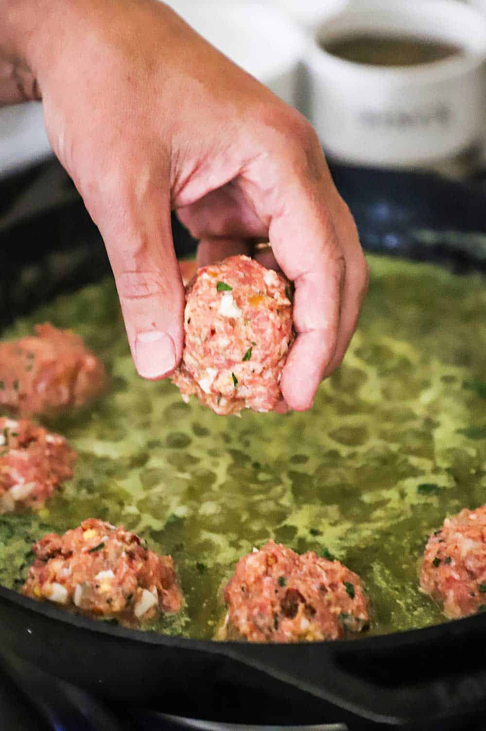 A person's hand placing an uncooked meatball into a skillet of simmering salsa verde.