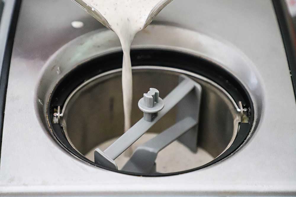 Blueberry ice cream custard being poured from a large measuring cup into an electric ice cream maker.