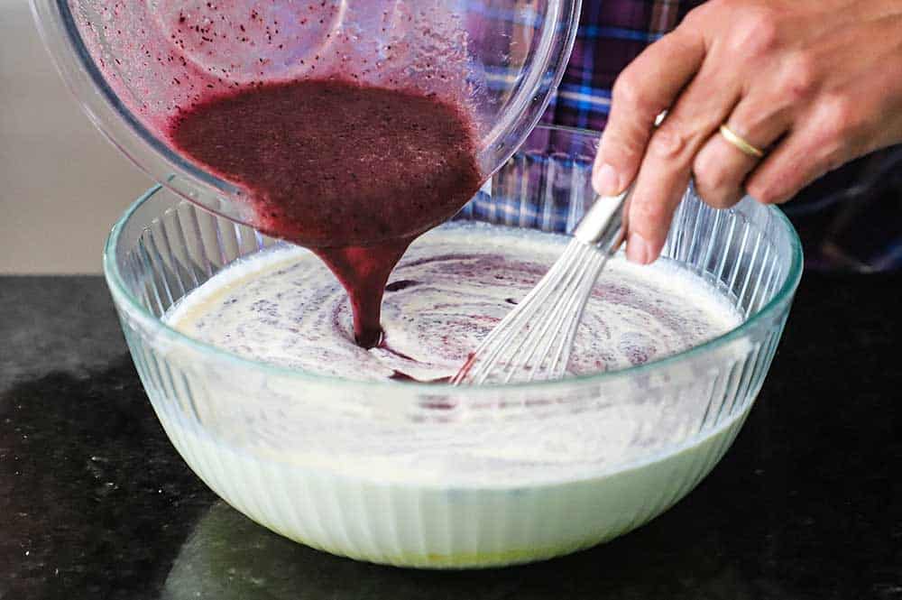 A person pouring puréed blueberries into a large glass bowl filled with ice cream custard.