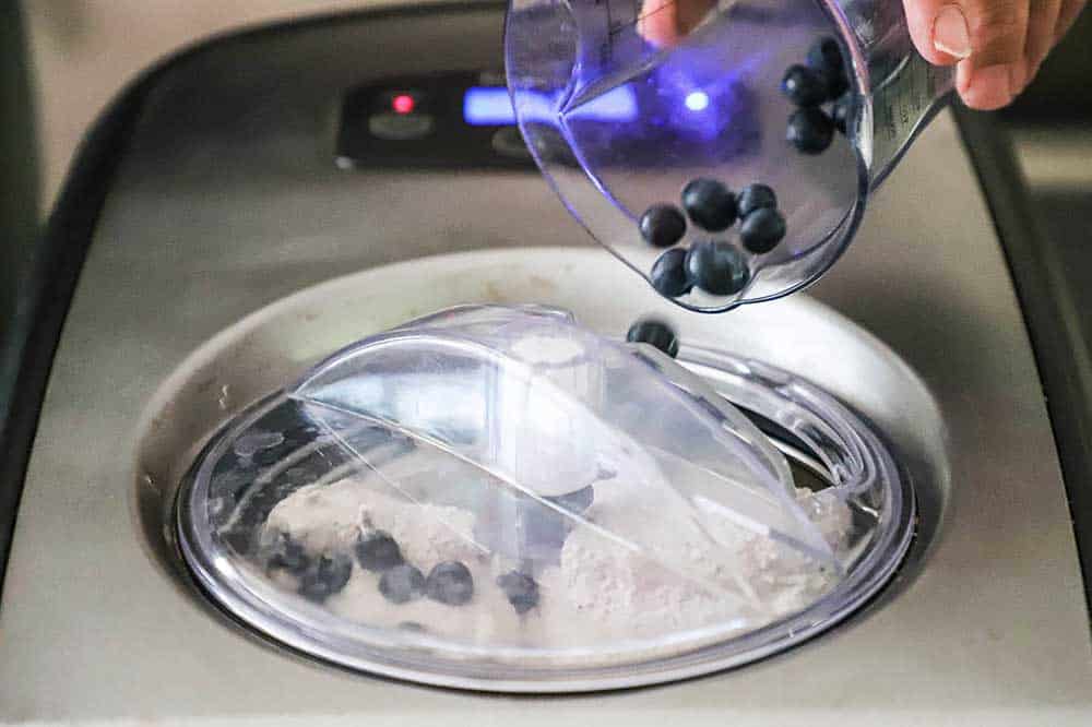 A person pouring fresh blueberries from a plastic measuring cup through an opening over a churning bowl of ice cream in an ice cream maker.