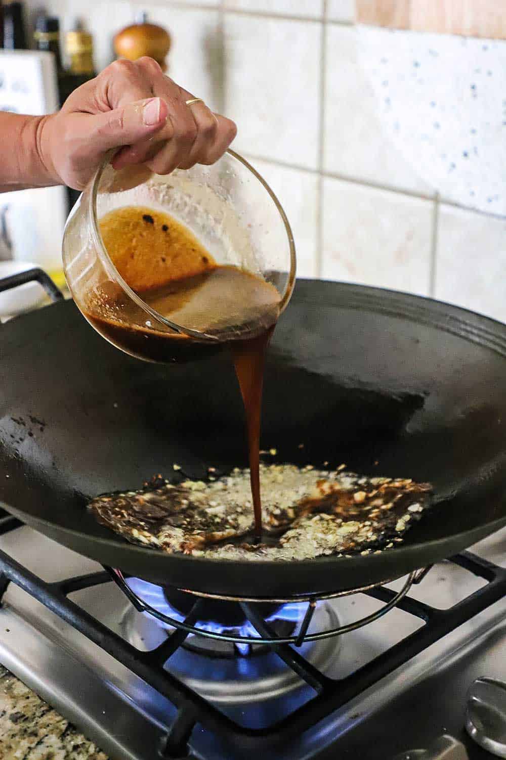 A person pouring a brown sauce into a wok filled with oil and garlic and ginger.